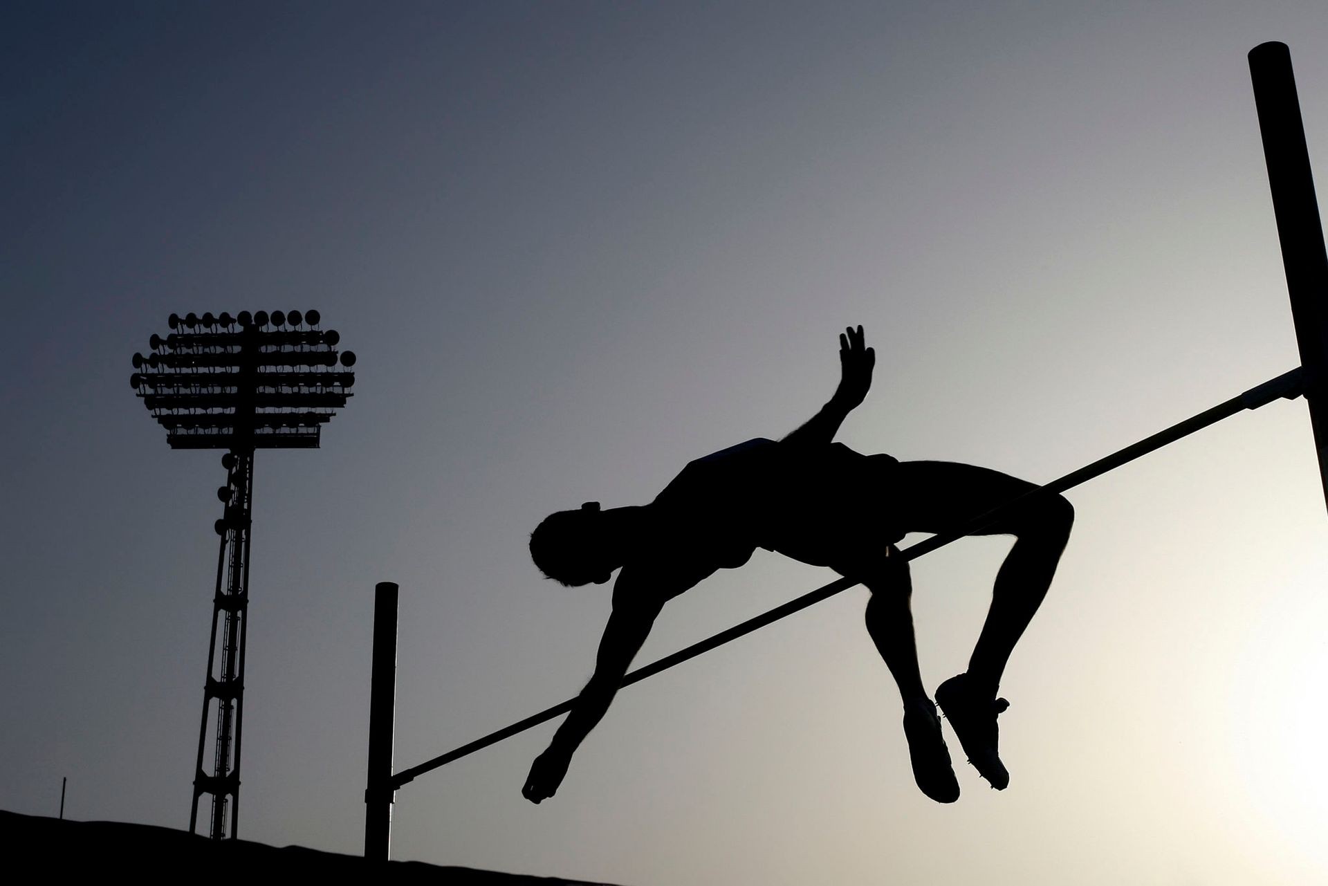 Silhouette of a high jumper clearing the bar with stadium lights in the background at dusk.