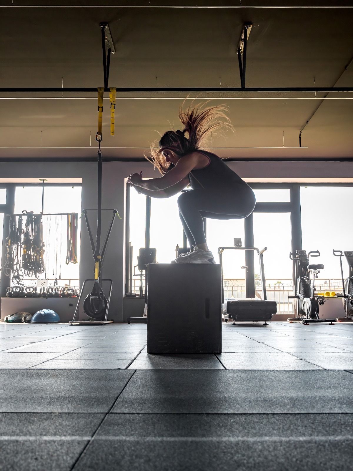 Person performing a box jump in a gym with fitness equipment and large windows in the background.